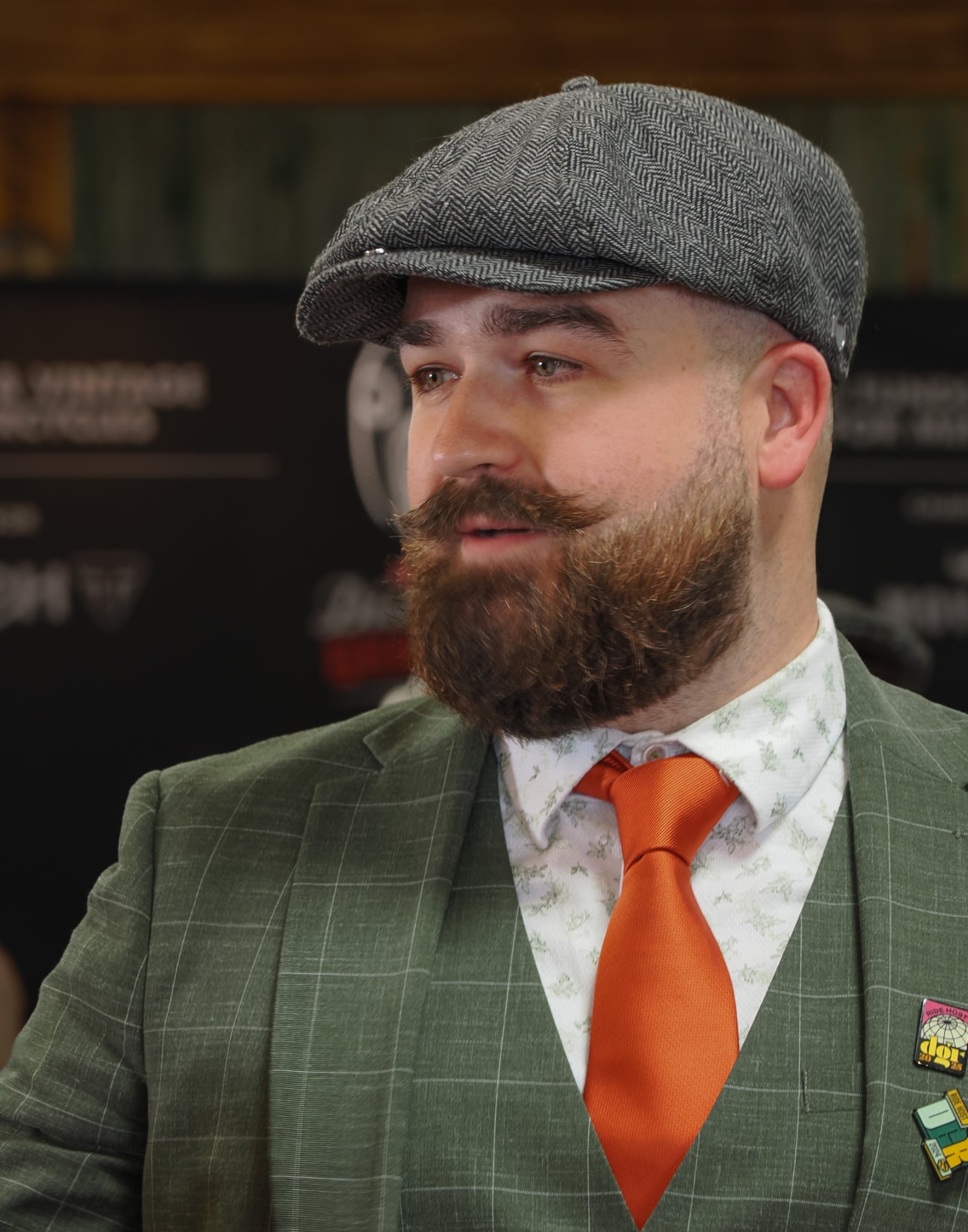 The image features a light-skinned man with a full beard and mustache, wearing a gray herringbone newsboy cap, a green plaid suit jacket, a patterned shirt, and a striking orange tie. He has "Ride Host" and "Dor" lapel pins. The background is dark. The shot is a medium shot.