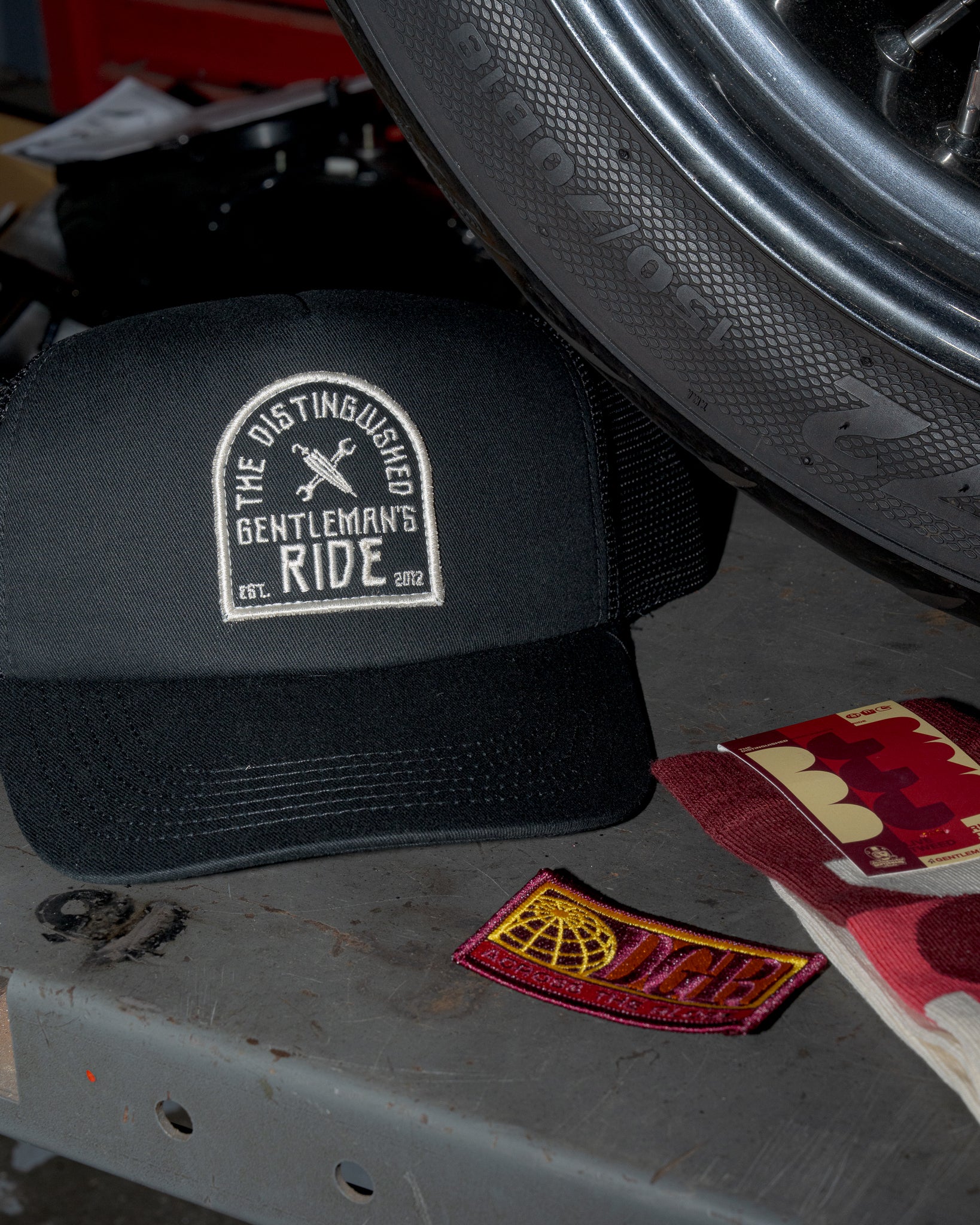 The image is a close-up shot featuring a black "The Distinguished Gentleman's Ride" baseball cap with a white embroidered logo, a motorcycle tire, a patch with a globe design and the letters "DGR", and socks in a red, white, and tan pattern on a gray workbench in what appears to be a garage or workshop setting. The black and white contrasts of the cap and the red sock and patch stand out in the scene.