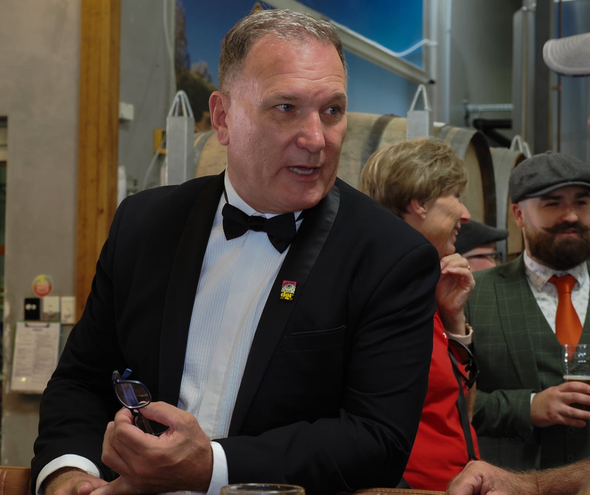The image captures a social gathering at a brewery, with the main subject being a man in a black tuxedo speaking while holding glasses. Behind him are other attendees including a smiling woman and a bearded man in a green suit and orange tie. The man in the tuxedo has a "One More dOR 2023" pin on his lapel. The scene is set against a backdrop of barrels and brewery equipment, suggesting a celebratory or event atmosphere.