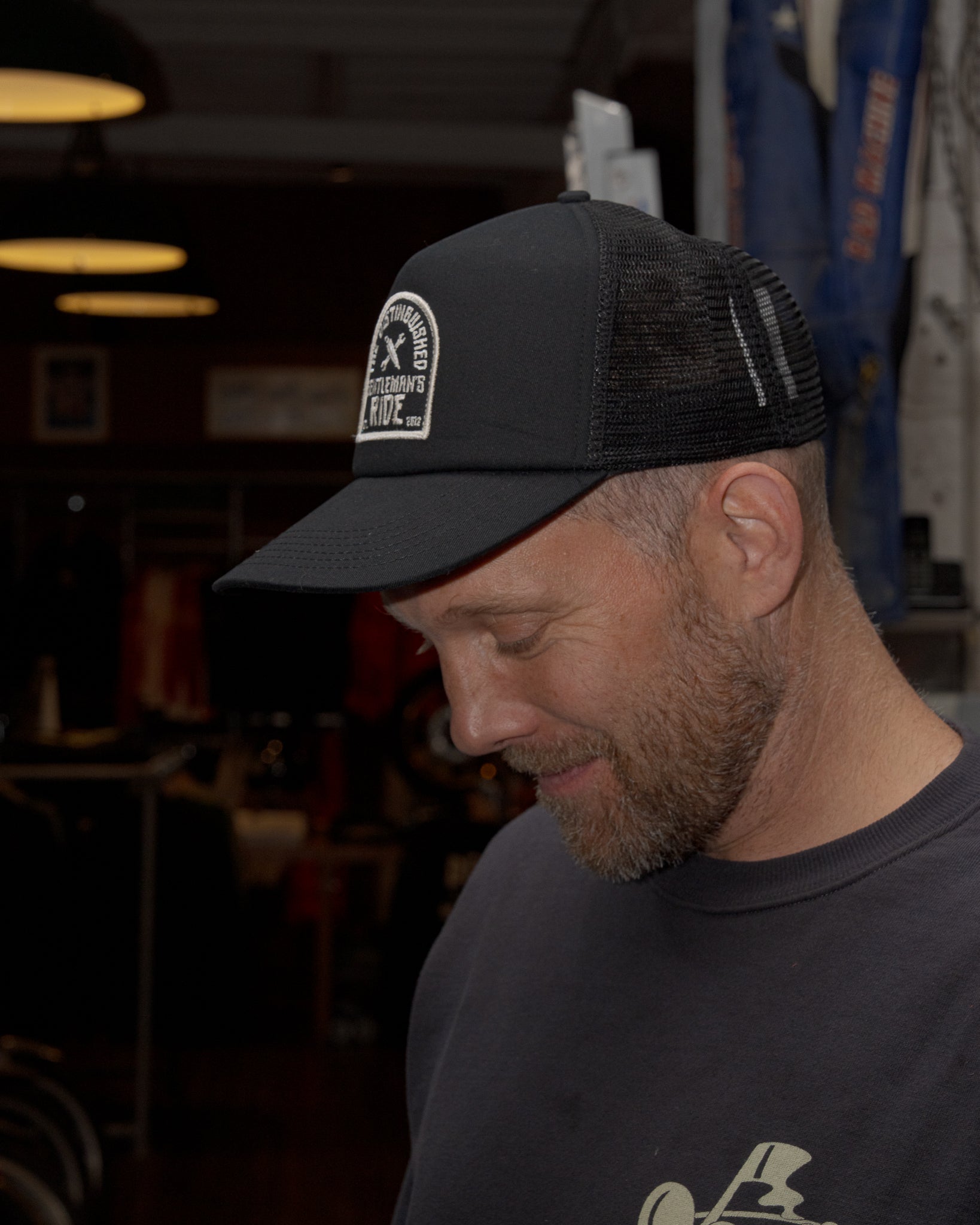 The image shows a man wearing a black trucker hat with a "Distinguished Gentleman's Ride 2012" logo patch. He is looking down with a soft smile, suggesting a contemplative or content mood. The scene is set indoors, possibly in a garage or workshop, with a blurred background and warm lighting. The composition focuses on the man's face and the hat, highlighting the logo.