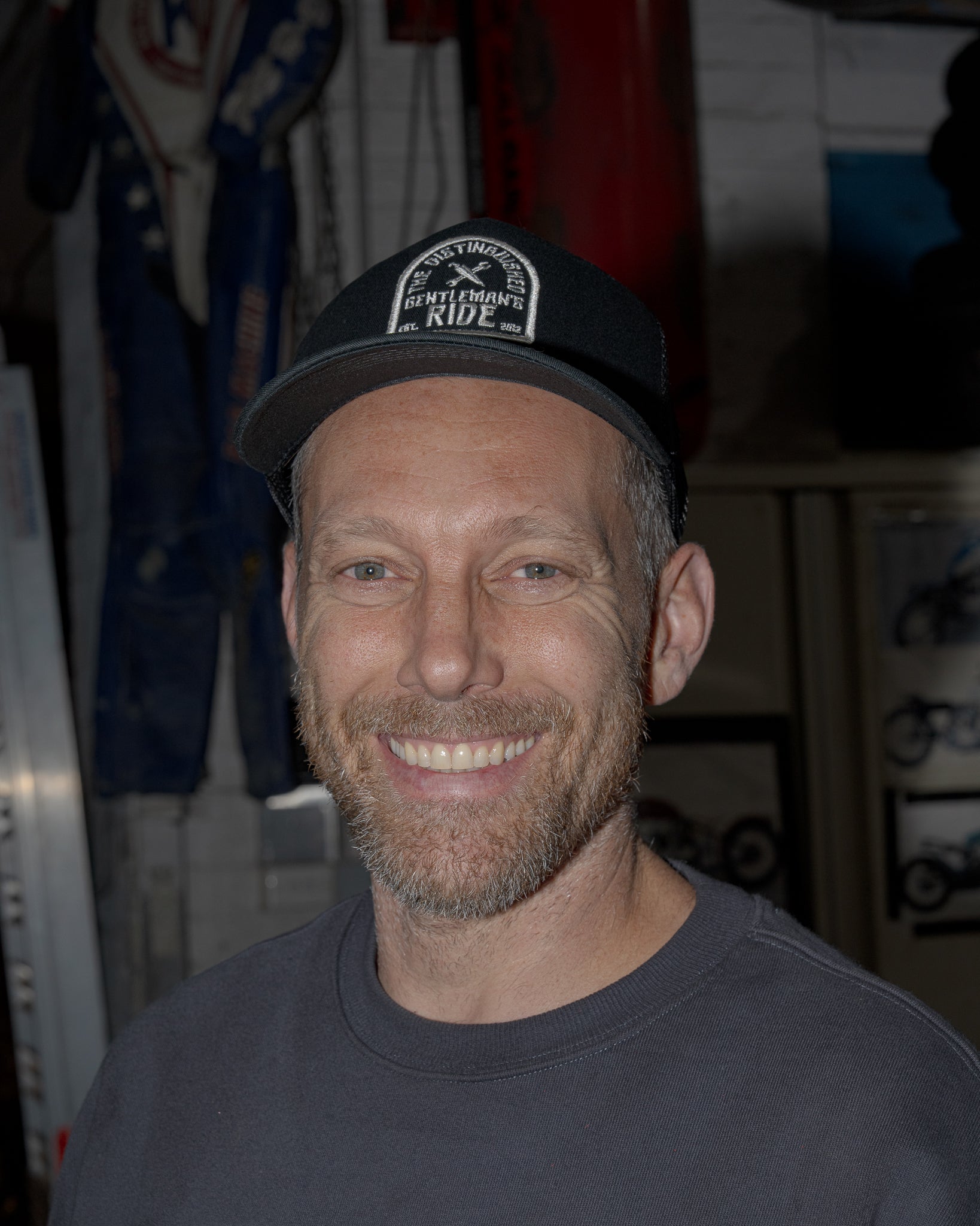 The image is a medium close-up of a smiling man wearing a gray t-shirt and a black baseball cap with "The Distinguished Gentleman's Ride Est. 2012" logo. The background is blurred, suggesting an indoor setting like a garage or workshop, with racing-related items visible. The man's expression and the logo on the cap indicate a cheerful and potentially charitable theme associated with the Distinguished Gentleman's Ride event.