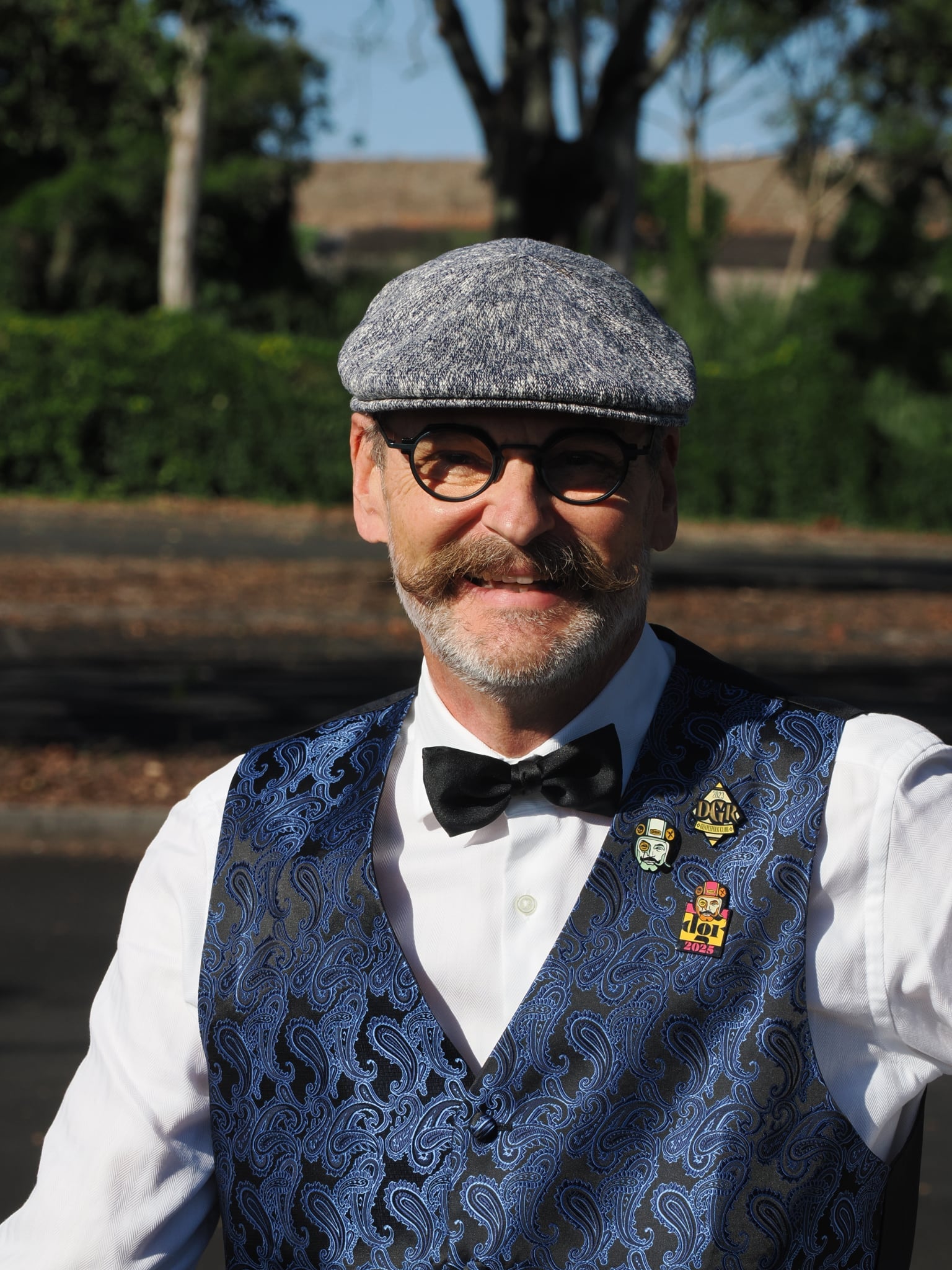 The image features a smiling middle-aged man wearing a gray tweed cap, round glasses, a black bow tie, and a blue paisley vest over a white shirt. He is standing outdoors in front of a blurred background of trees and a building. Several pins are affixed to his vest, including one that reads "2023 DGHO DOUALITIER KLUB." The composition is a medium shot focusing on the man's face and upper body, conveying a sense of happiness and formality.