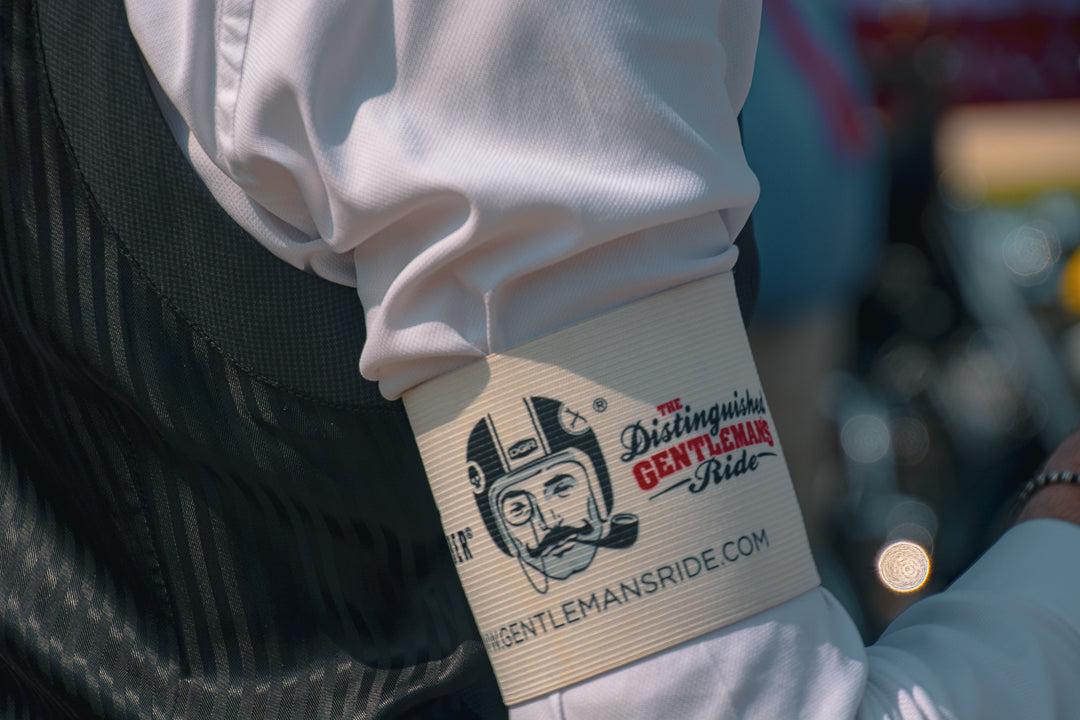 This close-up image features a participant in 'The Distinguished Gentleman's Ride,' an annual global event supporting men's mental health and prostate cancer research. The focus is on an arm adorned with a band displaying the event's logo: a dapper gentleman in a vintage motorcycle helmet with a monocle and pipe. The band also shows the event's name and website. The person is dressed in a white shirt and a dark, striped vest, indicating a focus on classic and stylish attire typical of the event. The blur...