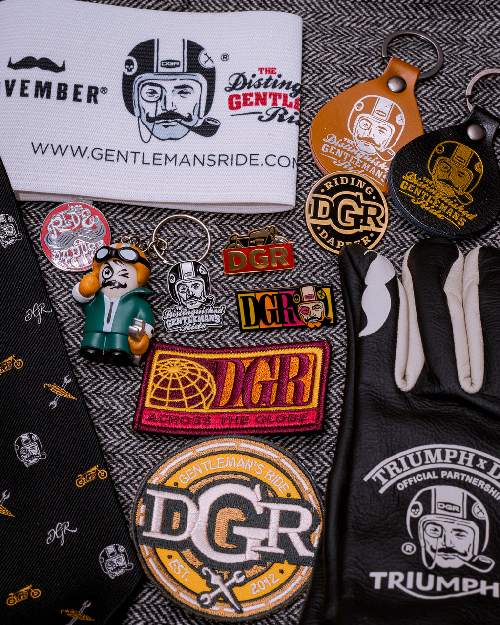 The image displays a collection of items related to the Distinguished Gentleman's Ride (DGR), including branded pins, patches, keychains, a tie, and Triumph-branded leather gloves. The arrangement is set against a gray and white herringbone background. The merchandise features the DGR logo, depicting a stylized man with a mustache, monocle, and helmet, and promotional text such as "NOVEMBER," "WWW.GENTLEMANSRIDE.COM," and "RIDING DGR DAPPER." The color scheme includes black, white, gold, brown, red, gree...