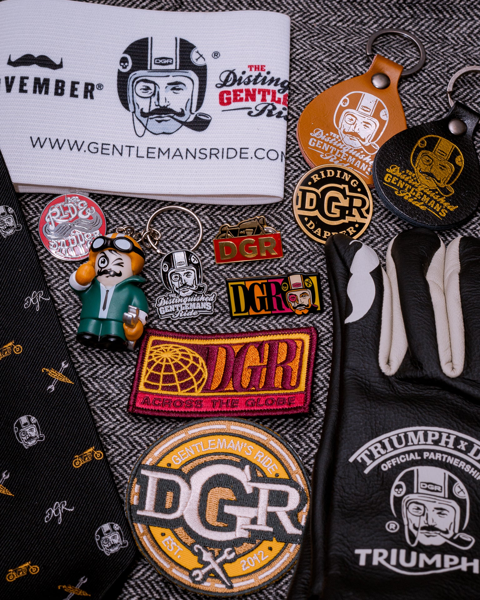 The image displays a collection of items related to the Distinguished Gentleman's Ride (DGR), including branded pins, patches, keychains, a tie, and Triumph-branded leather gloves. The arrangement is set against a gray and white herringbone background. The merchandise features the DGR logo, depicting a stylized man with a mustache, monocle, and helmet, and promotional text such as "NOVEMBER," "WWW.GENTLEMANSRIDE.COM," and "RIDING DGR DAPPER." The color scheme includes black, white, gold, brown, red, gree...