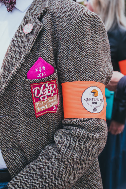 The image is a close-up shot detailing the attire of a participant in an event such as the Distinguished Gentleman's Ride. The subject is wearing a tweed jacket, a white shirt, and a burgundy bow tie, accessorized with a pink "DGR 2016" handkerchief, a DGR patch that reads "Live Tweed & Ride Dapper," and an orange armband with the "Gentleman Lignano Sabbiadoro FVG" logo. The focus is on the texture of the tweed and the distinct branding associated with the event.
