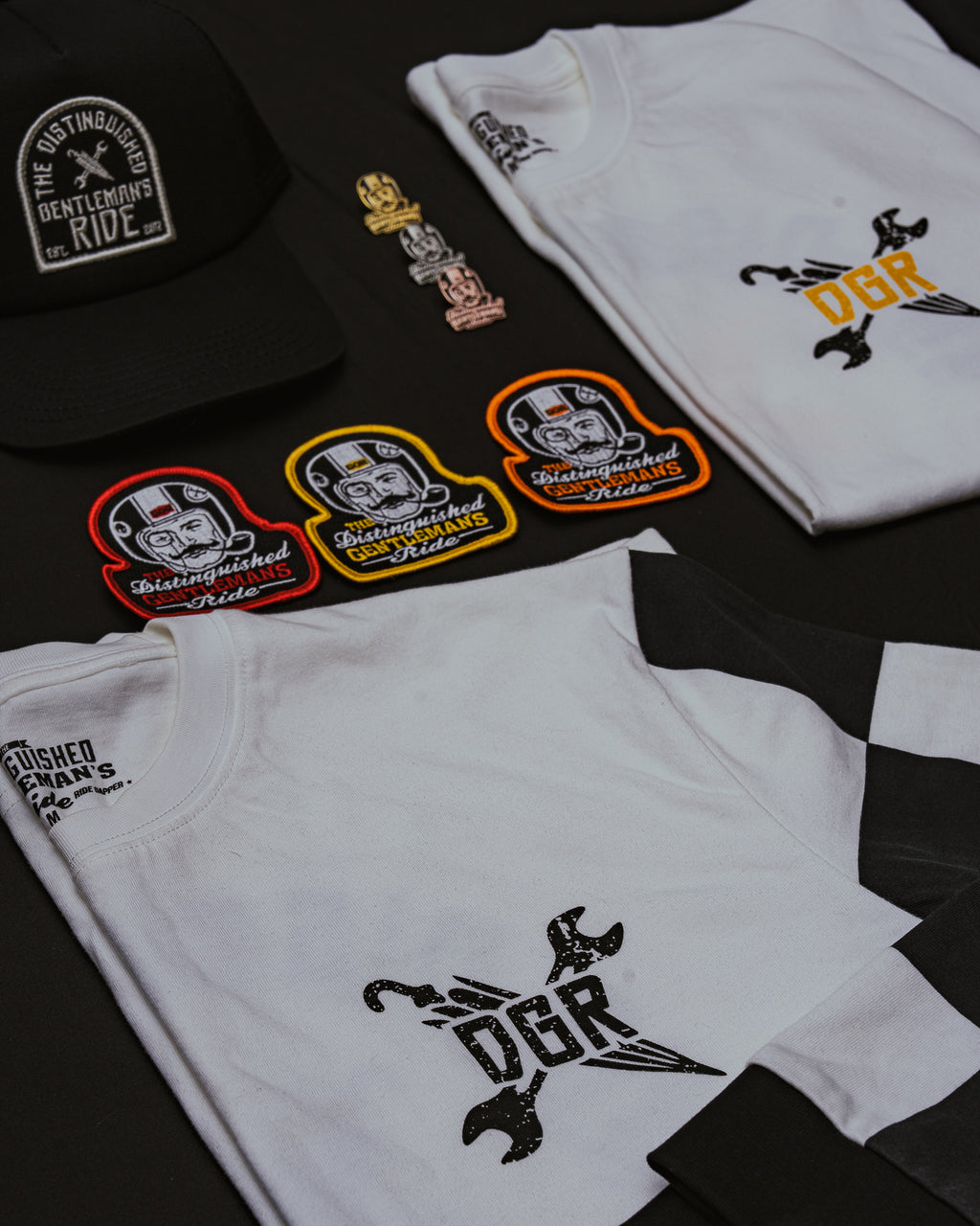The image showcases merchandise related to "The Distinguished Gentleman's Ride," including a black baseball cap, three enamel pins, three iron-on patches, and two white T-shirts with the "DGR" logo. The items are laid out on a black surface. The merchandise features the event's name and logo, emphasizing the theme of the dapper style and community associated with the ride.