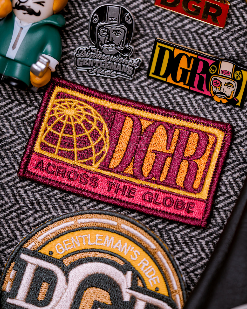 The image features a close-up of various patches and pins related to "The Distinguished Gentleman's Ride" (DGR). Predominant colors include maroon, yellow, gold, and black, set against a herringbone background. The items include a rectangular patch with a globe and "DGR ACROSS THE GLOBE" text, several DGR-branded pins, and a partial view of a "GENTLEMAN'S RIDE" patch. A plastic toy figurine is also present. The composition and lighting suggest a deliberate arrangement, possibly for promotional material.