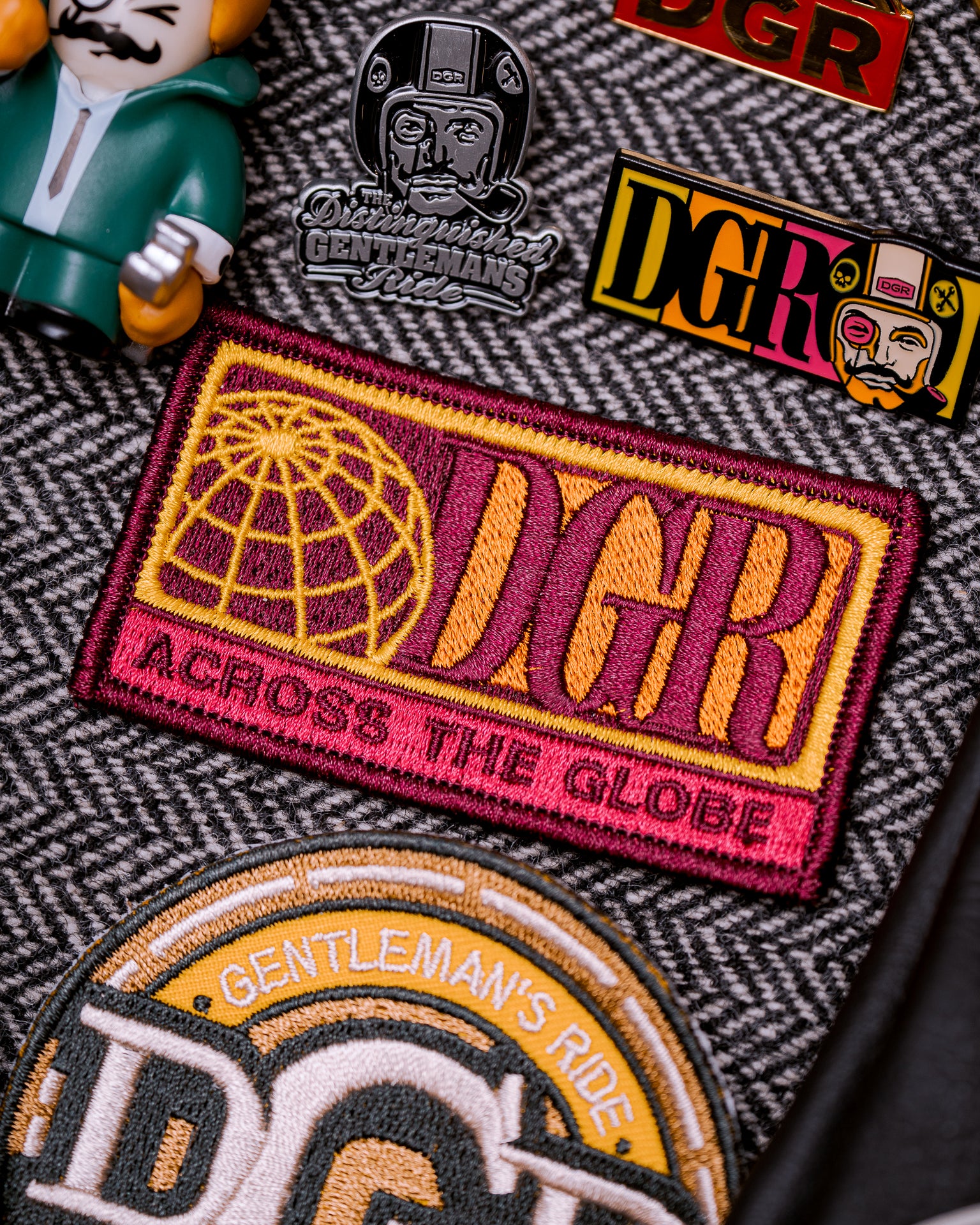 The image features a close-up of various patches and pins related to "The Distinguished Gentleman's Ride" (DGR). Predominant colors include maroon, yellow, gold, and black, set against a herringbone background. The items include a rectangular patch with a globe and "DGR ACROSS THE GLOBE" text, several DGR-branded pins, and a partial view of a "GENTLEMAN'S RIDE" patch. A plastic toy figurine is also present. The composition and lighting suggest a deliberate arrangement, possibly for promotional material.